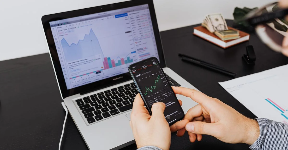 Personne analysant des données boursières sur un ordinateur portable et un smartphone à l'intérieur.