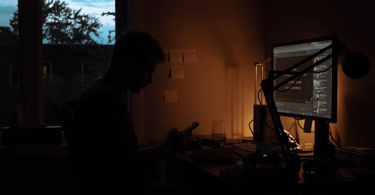 Silhouette d'un homme utilisant un smartphone dans une pièce sombre éclairée par l'écran d'ordinateur.