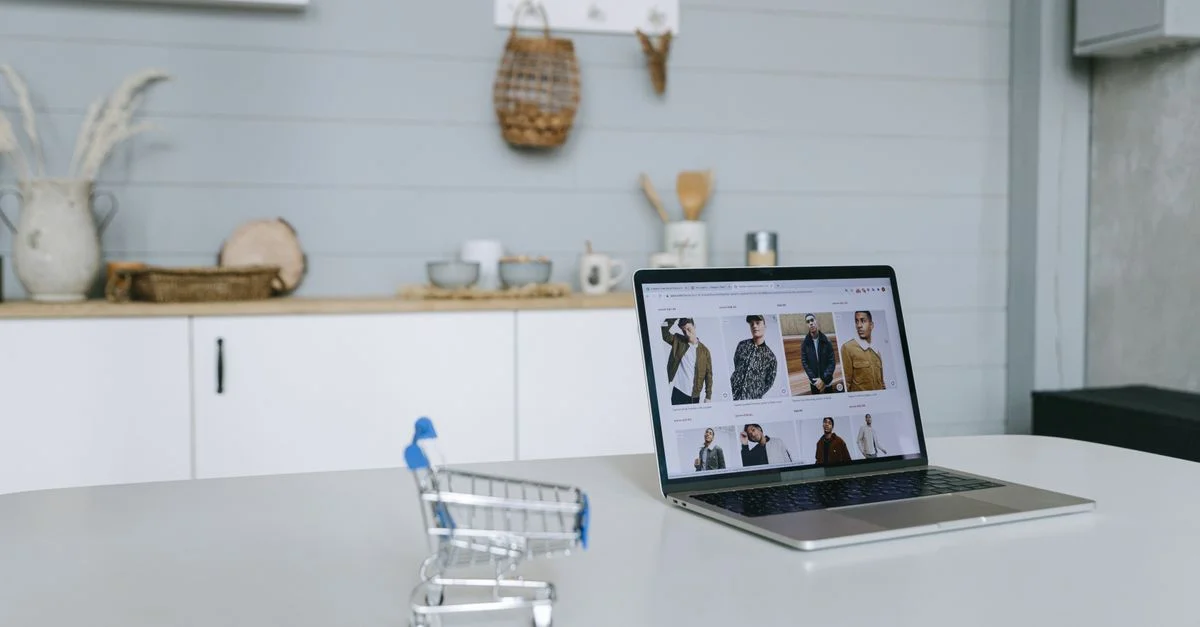 Un ordinateur portable affichant un portail de shopping en ligne est posé sur un bureau minimal avec un chariot de shopping miniature en focus.