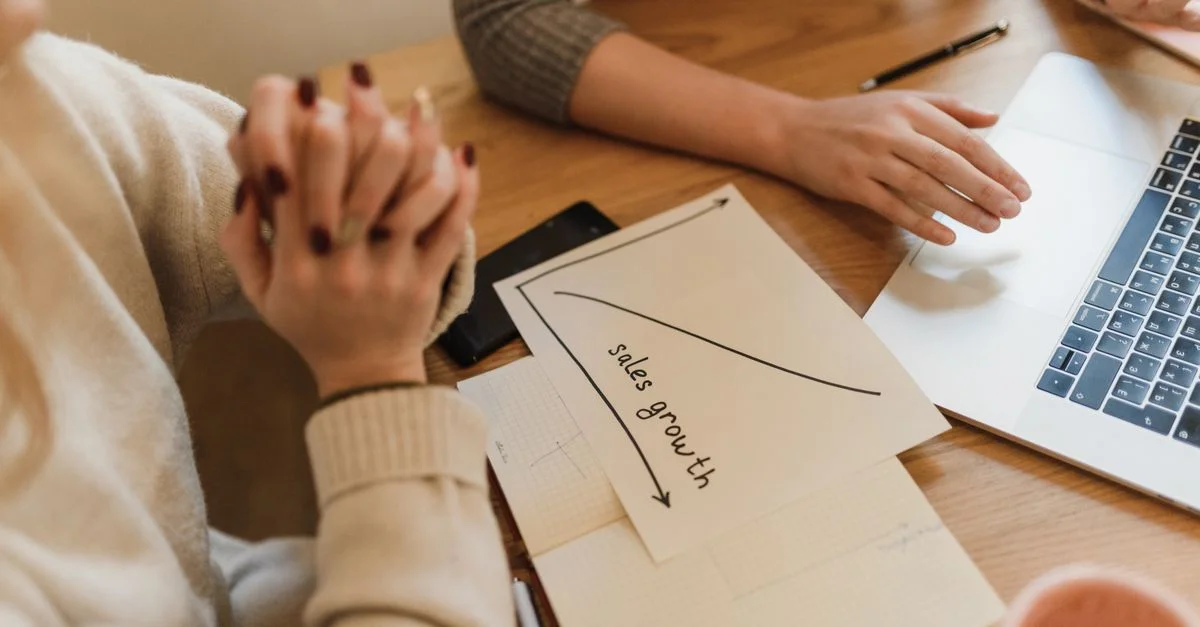 Deux femmes collaborent à un bureau en discutant de la croissance des ventes avec un ordinateur portable et des notes.