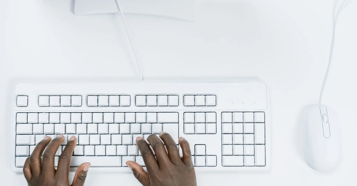 Vue de dessus des mains tapant sur un clavier d'ordinateur blanc et une souris, sur un bureau blanc.